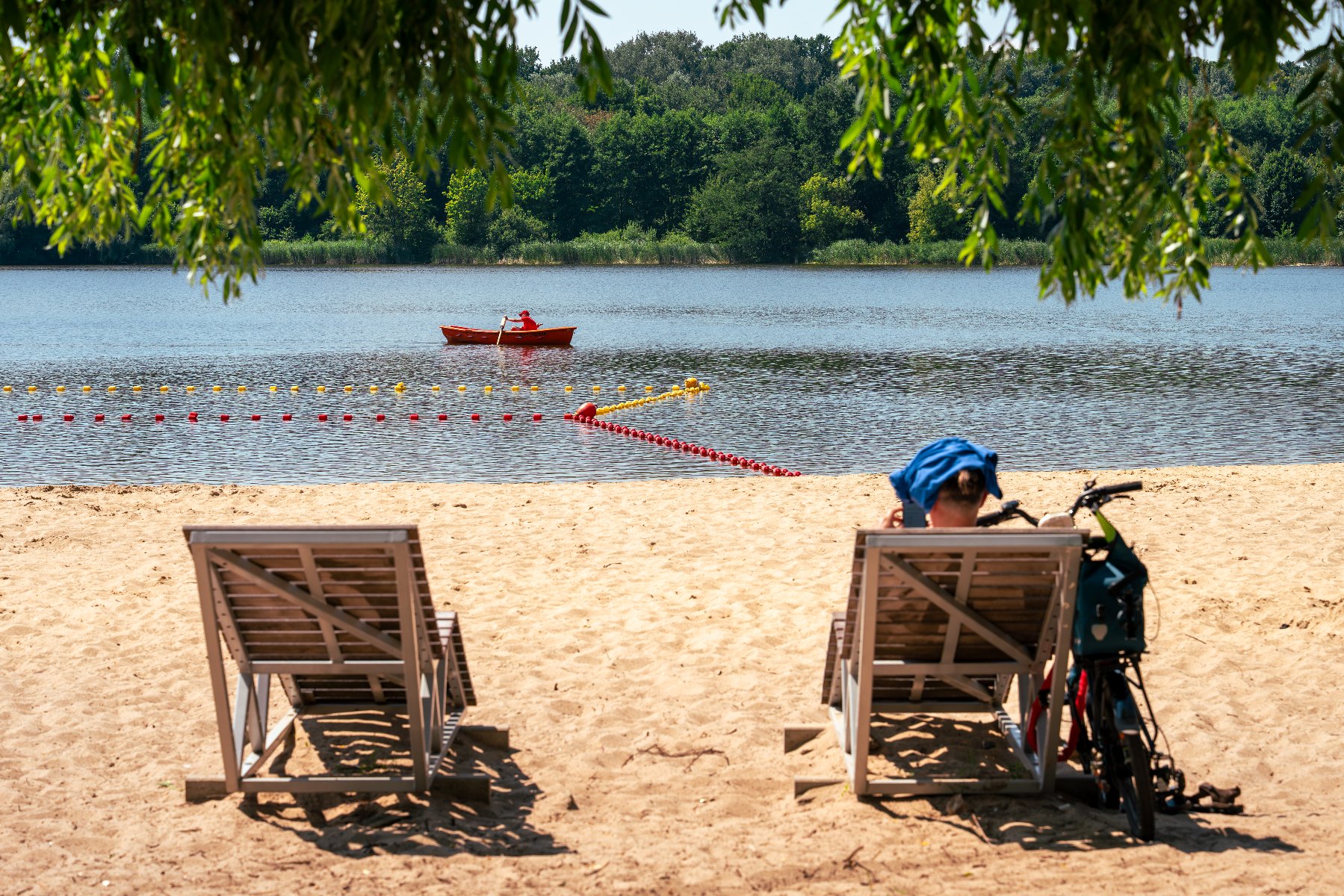 Osoba wypoczywająca na leżaku na piaszczystej plaży nad jeziorem, z widokiem na wyznaczone kąpielisko i łódź ratunkową na wodzie.