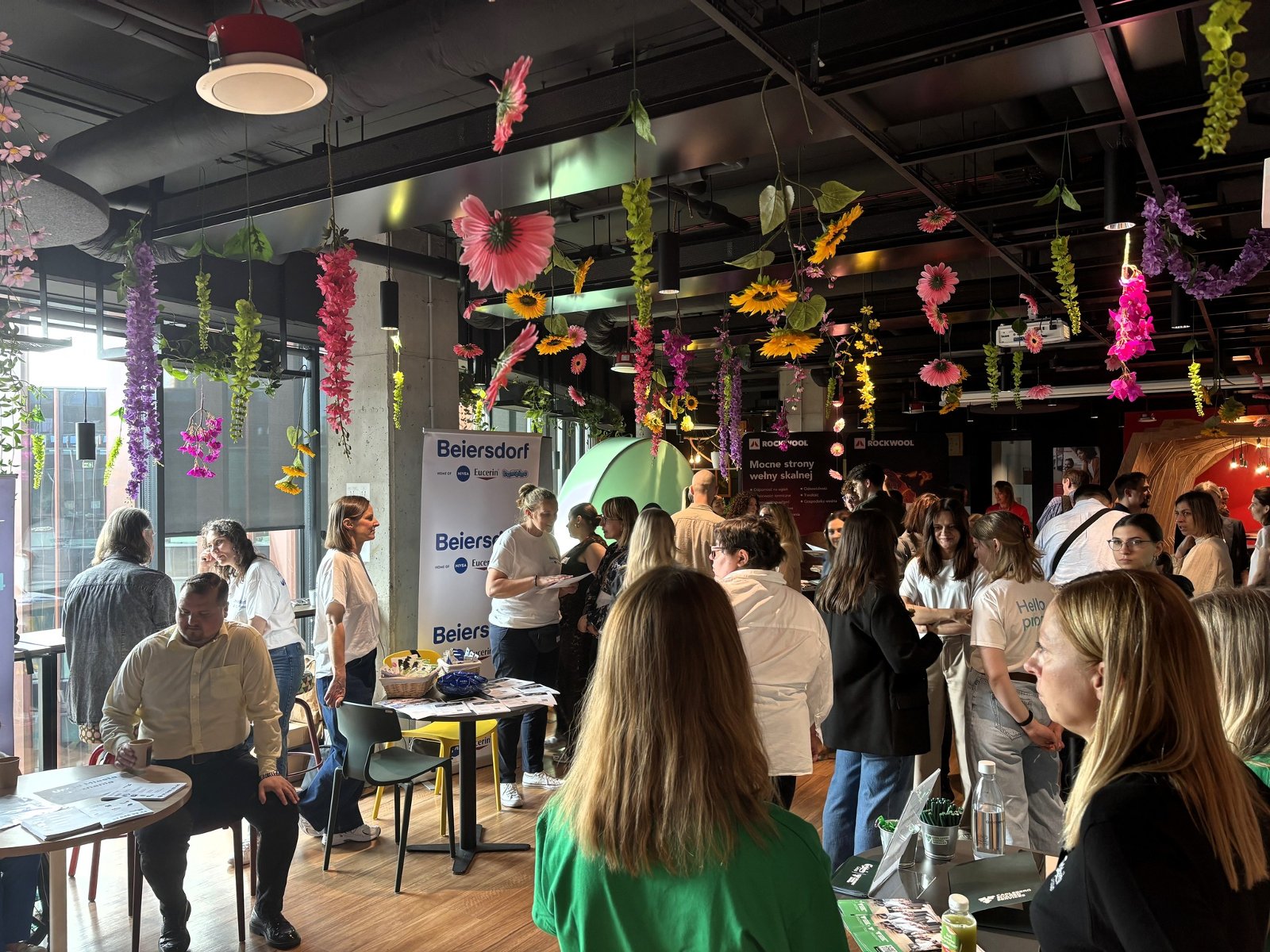 Otwarta przestrzeń biurowa. Przy rozłożonych stoiskach prezentują się różne firmy. Mają ulotki, gadżety, informatory. Przestrzeń wypełniają ludzie. Rozmawiają, spacerują.