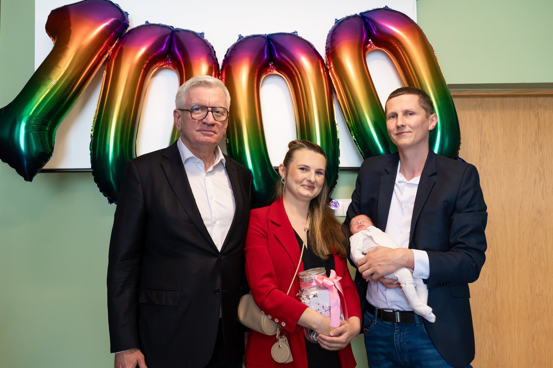 Pamiątkowe zdjęcie grupowe Prezydenta Miasta Poznania Jacka Jaśkowiaka z rodzicami trzymającymi noworodka na tle tęczowych balonów tworzących liczbę 1000.