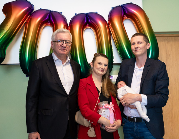 Pamiątkowe zdjęcie grupowe Prezydenta Miasta Poznania Jacka Jaśkowiaka z rodzicami trzymającymi noworodka na tle tęczowych balonów tworzących liczbę 1000.