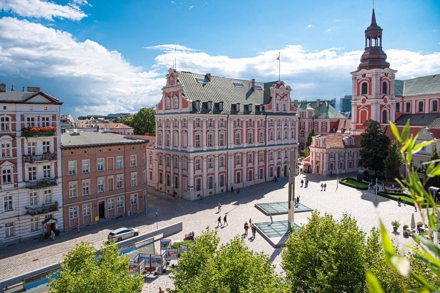 Panorama placu Kolegiackiego w Poznaniu. W centrum kadru dominuje zabytkowy, różowo-biały gmach Urzędu Miasta (dawne Kolegium Jezuickie). Po prawej stronie widać wieżę bazyliki farnej. Plac jest wybrukowany, widać na nim spacerujących ludzi, nowoczesną fontannę oraz wystawę plenerową.