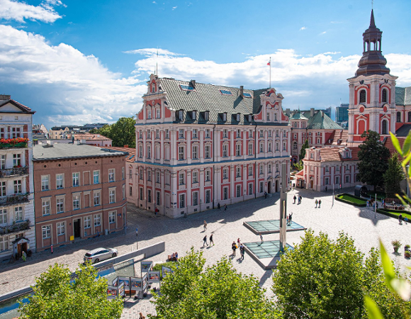 Panorama placu Kolegiackiego w Poznaniu. W centrum kadru dominuje zabytkowy, różowo-biały gmach Urzędu Miasta (dawne Kolegium Jezuickie). Po prawej stronie widać wieżę bazyliki farnej. Plac jest wybrukowany, widać na nim spacerujących ludzi, nowoczesną fontannę oraz wystawę plenerową.