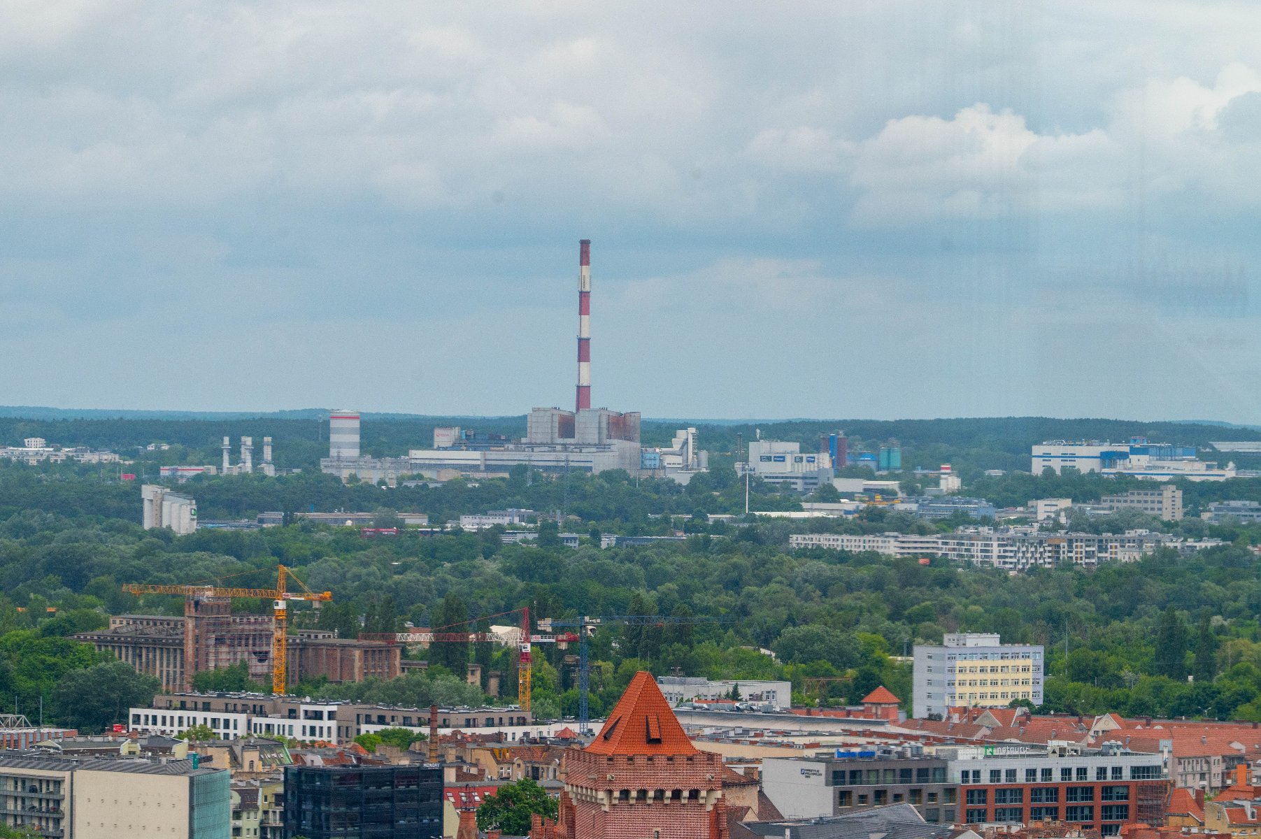 Panorama Poznania. Na pierwszym planie dachy budynków i szczyt wieży kościelnej. W tle widoczny jest las oraz zabudowania miejskie, w tym wysoki, biało-czerwony komin elektrociepłowni.