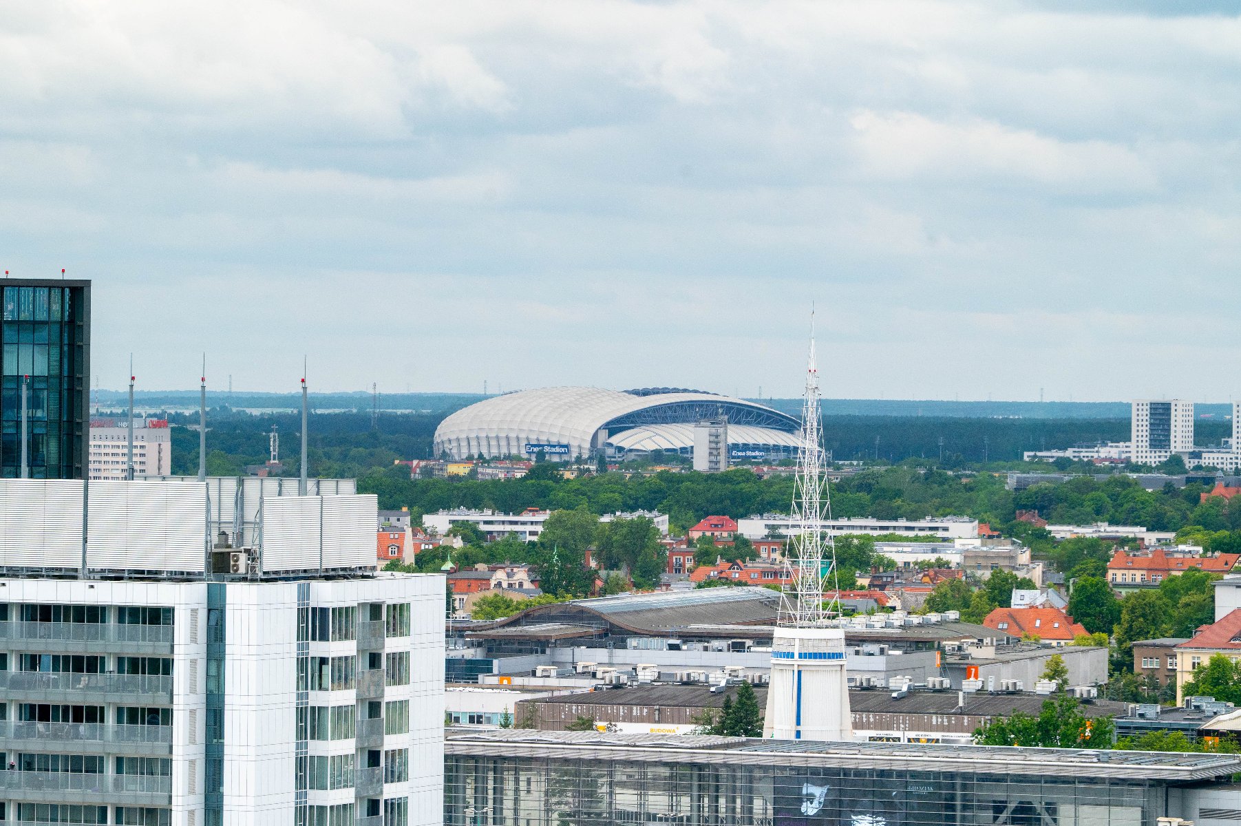 Panoromiczny widok na miejską zabudowę z lotu ptaka, z widoczną w tle białą czaszą stadionu Enea Stadion oraz jasną wieżą iglicową na tle pochmurnego nieba.