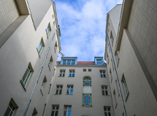 Perspektywiczne ujęcie z dołu przedstawiające wąski dziedziniec otoczony wysokimi, jasnymi ścianami kamienic. W centralnej części widać fasadę z ozdobnymi, witrażowymi oknami o zielonych i niebieskich szybkach. Powyżej znajduje się fragment dachu z czerwoną dachówką i lukarnami, a nad całością góruje błękitne niebo z delikatnymi chmurami.