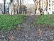 Piaszczysto-trawiasta ścieżka na lekkim wzniesieniu. Po bokach widoczne są pomarańczowe znaczniki wbijane w ziemię. Na wzniesieniu w oddali widoczna jest biała furgonetka i ogrodzenie z drewnianych pali. W tle widoczne są bloki mieszkalne z białymi fasadami i gołe drzewa.