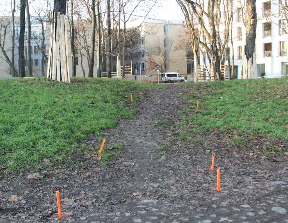 Piaszczysto-trawiasta ścieżka na lekkim wzniesieniu. Po bokach widoczne są pomarańczowe znaczniki wbijane w ziemię. Na wzniesieniu w oddali widoczna jest biała furgonetka i ogrodzenie z drewnianych pali. W tle widoczne są bloki mieszkalne z białymi fasadami i gołe drzewa.