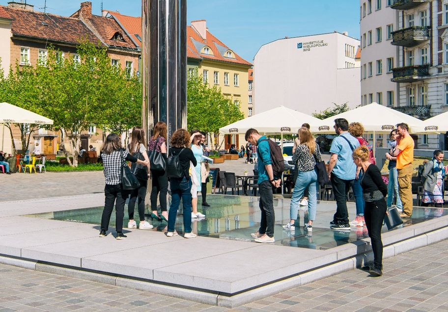 plac z ogródkami gastronomicznymi, wokół kamienice, w posadzce placu ekspozycja za szklanymi szybami, ogląda ją grupa osób patrząc w dół, nad espopzycją wznosi się stella z lustrzaną powiechrznią