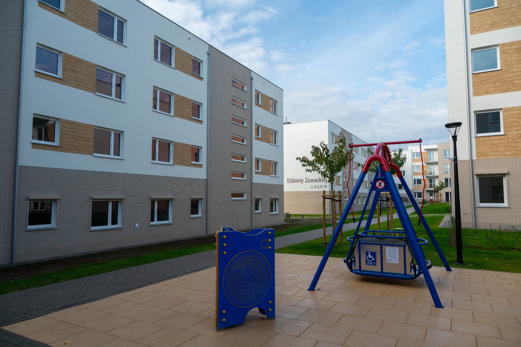 Plac zabaw z czerwoną i niebieską huśtawką przystosowaną dla osób z niepełnosprawnościami na tle nowoczesnych, jasnych budynków mieszkalnych. Na jednym z budynków widoczny napis: "Elżbiety Zawackiej". Po lewej stronie niebieski panel edukacyjny.