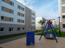 Plac zabaw z czerwoną i niebieską huśtawką przystosowaną dla osób z niepełnosprawnościami na tle nowoczesnych, jasnych budynków mieszkalnych. Na jednym z budynków widoczny napis: "Elżbiety Zawackiej". Po lewej stronie niebieski panel edukacyjny.