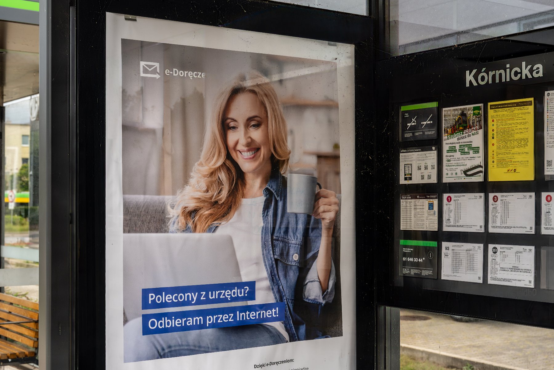 Plakat reklamowy usługi e-Doręczenia na przystanku tramwajowym Kórnicka, przedstawiający uśmiechniętą kobietę z laptopem i kubkiem.