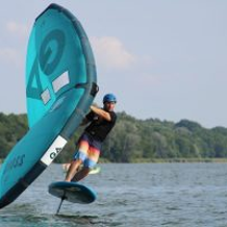 Zdjęcie osoby uprawiającej wingfoiling na jeziorze. Sportowiec, ubrany w kask i kolorowe spodenki, unosi się nad powierzchnią wody na desce. W dłoniach trzyma duży, turkusowy żagiel z ciemnymi detalami, który jest wygięty pod wpływem wiatru. W tle widać gęstą linię zielonych drzew na brzegu oraz jasne, pogodne niebo.