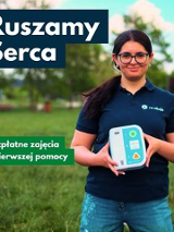 Kobieta stojąca na tle trawy i drzew trzyma automatyczny defibrylator AED. Ma okulary i granatową koszulkę z logo. Na plakacie widnieją napisy: "Ruszamy Serca" oraz "Bezpłatne zajęcia z pierwszej pomocy".
