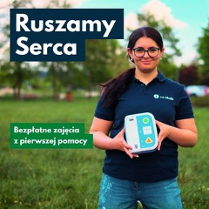 Kobieta stojąca na tle trawy i drzew trzyma automatyczny defibrylator AED. Ma okulary i granatową koszulkę z logo. Na plakacie widnieją napisy: "Ruszamy Serca" oraz "Bezpłatne zajęcia z pierwszej pomocy".
