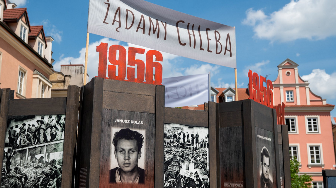 Plenerowa instalacja upamiętniająca wydarzenia z 1956 roku. Na pierwszym planie duże czerwone cyfry "1956", nad nimi biały baner z napisem "Żądamy chleba". Poniżej znajdują się czarno‑białe zdjęcia uczestników tamtych wydarzeń. W tle pastelowe kamienice z czerwonymi dachami i częściowo zachmurzone niebo.