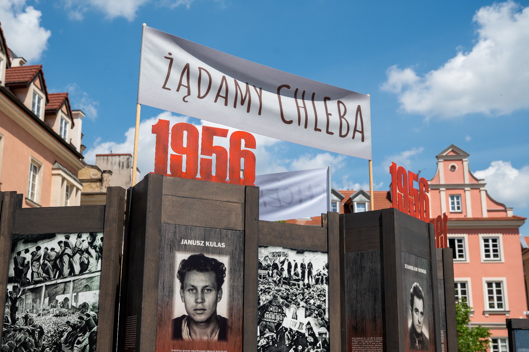 Plenerowa wystawa historyczna poświęcona Poznańskiemu Czerwcowi 1956 roku, wyeksponowana na tle miejskiej zabudowy z kamienicami o pastelowych elewacjach. Na ciemnobrązowych, drewnianych pylonach umieszczone są czarno-białe fotografie archiwalne, w tym portret Janusza Kulasa oraz zdjęcia przedstawiające demonstracje i zgromadzone tłumy.