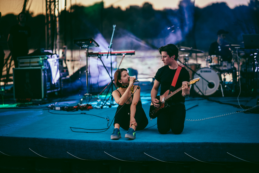 Plenerowy koncert muzyczny. Na scenie dwóch muzyków na pierwszym planie - jeden klęczy i gra na gitarze elektrycznej, drugi siedzi na podłodze sceny, trzyma mikrofon i śpiewa.