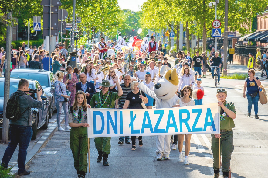 Pochód idących ulicą ludzi. Idący z przodu trzymają transparent z napisem "Dni Łazarza".