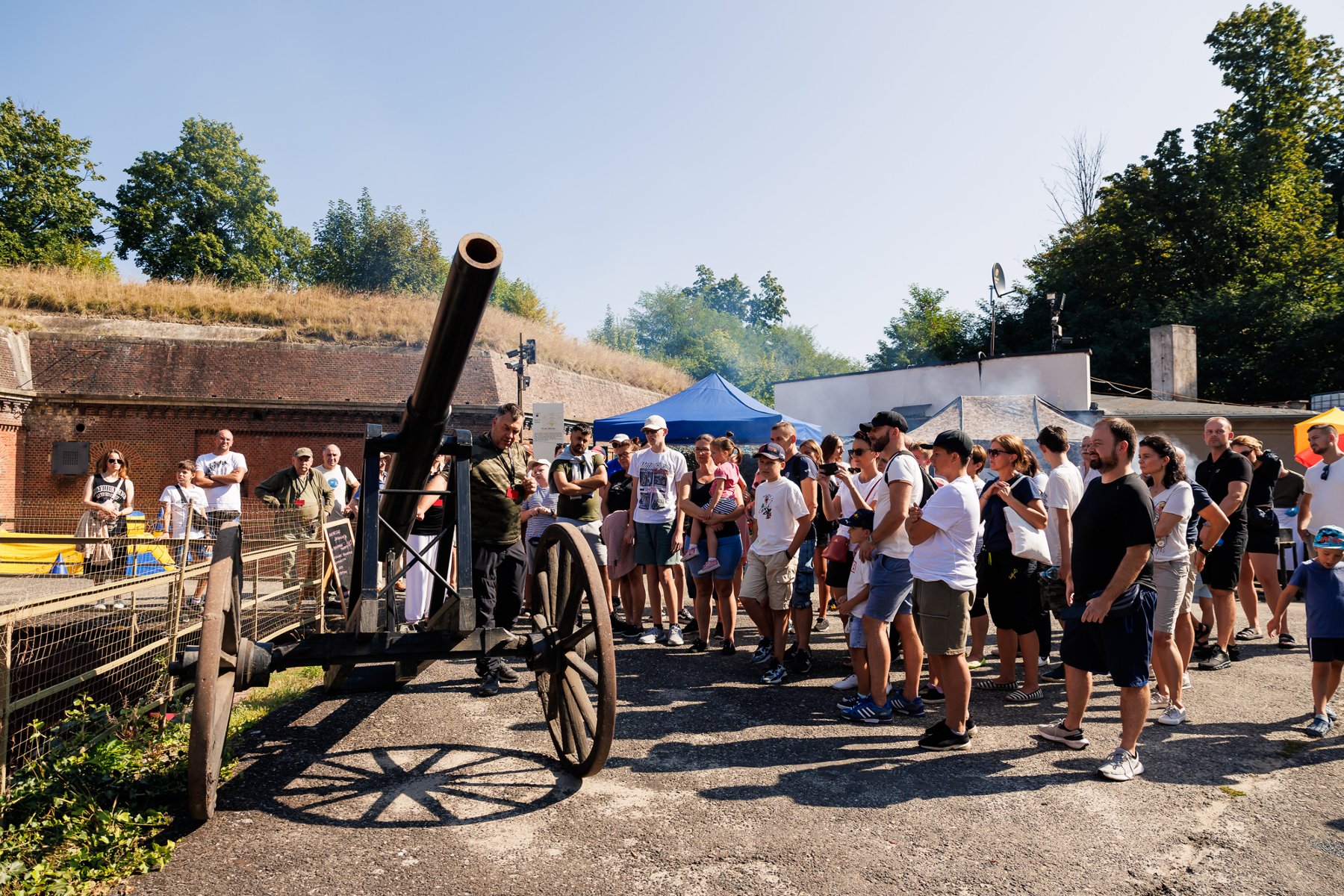 Pokaz historyczny z użyciem czarnej armaty na kołach, otoczony przez tłum odwiedzających w słoneczny dzień, na terenie ceglanej fortyfikacji.