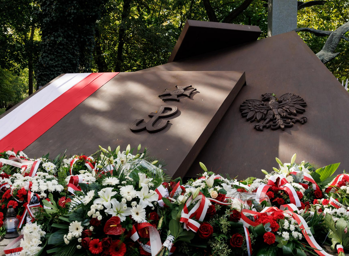 pomnik w kształcie nałożonych na siebie dużych płyt na nich wizerunki godła Polski symbolu harcerstwa i Polski Walczącej pod pomnikiem złożone wiązanki kwiatów i biało czerwona flaga
