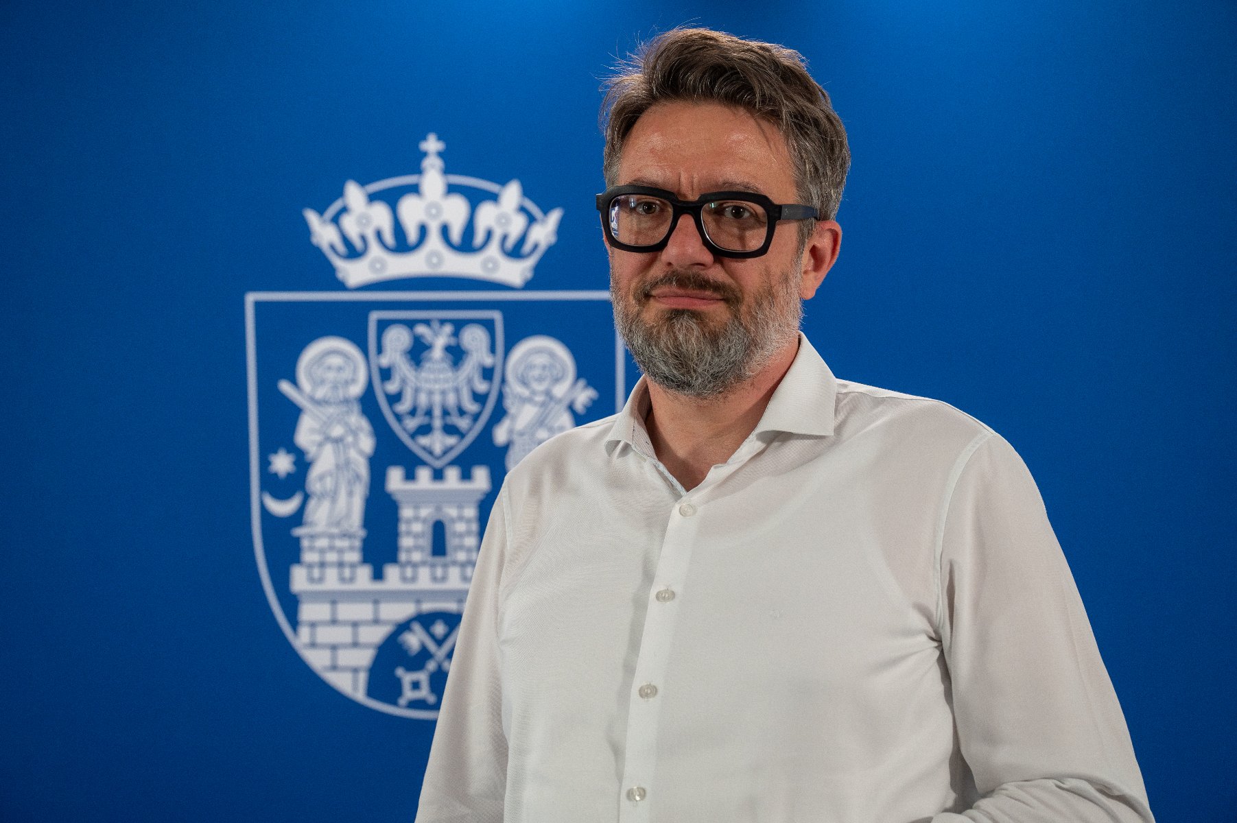 Portret mężczyzny w okularach z grubymi, czarnymi oprawkami i z zarostem, ubranego w białą koszulę. Stoi on na tle niebieskiej ścianki, na której widnieje biały herb Poznania.