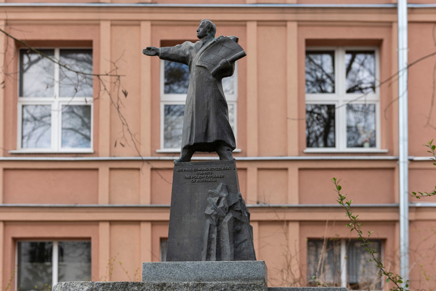 Posąg Sir Paula Edmunda Strzeleckiego, znanego jako 'Polski Odkrywca Australii.' Posąg przedstawia Strzeleckiego w długim płaszczu, z jedną ręką wyciągniętą do przodu, a drugą trzymającą książkę lub dokument. Posąg umieszczony jest na piedestale z napisem: 'SIR PAUL EDMUND STRZELECKI 1797-1873 POLSKI ODKRYWCA AUSTRALII.' W tle znajduje się budynek z brzoskwiniową fasadą i kilkoma oknami.