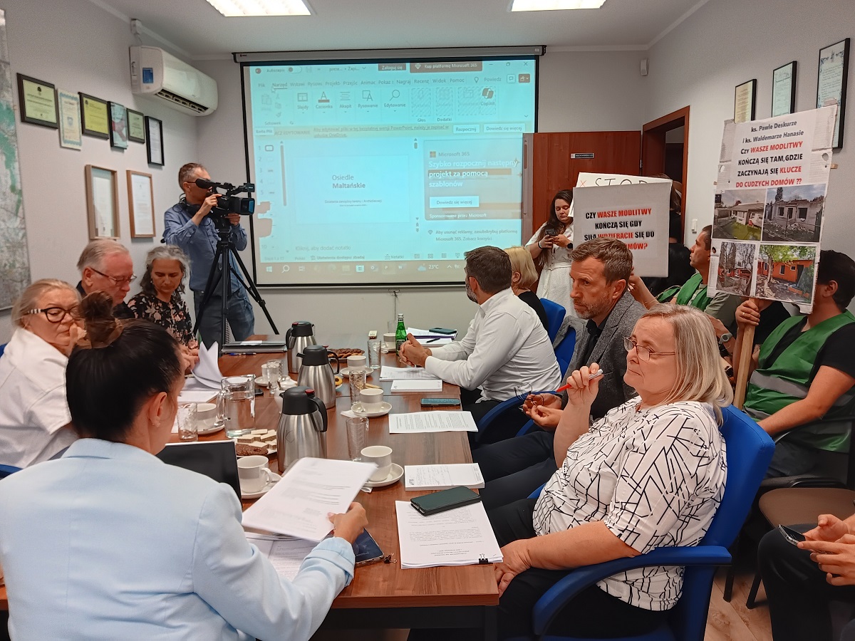 Posiedzenie Komisji Mieszkaniowej w dniu 10 września 2025 r., ludzie siedzą przy stole, niektórzy siedzą na krzesłach przy ścianie i trzymają transparenty