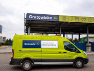 Profil jaskrawozielonego furgonu z napisem "Gratowóz" oraz adresem strony internetowej "poznan.pl/odpady" na boku. Samochód stoi na placu wyłożonym kostką brukową przed wiatą z dużym napisem "Gratowisko". Na dachu wiaty widoczne są piktogramy przedstawiające odpady, takie jak pralka, fotel czy lodówka. W tle widać kontenery oraz pochmurne niebo..