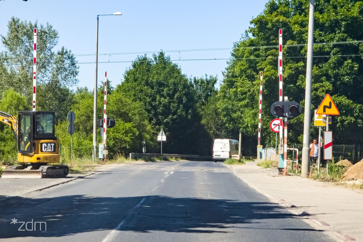 Przejazd kolejowy z otwartymi szlabanami w ciągu ul. Naramowickiej. Wzdłuż jezdni bariery ochronne i lampy. Na środku jezdni podwójna linia ciągła.