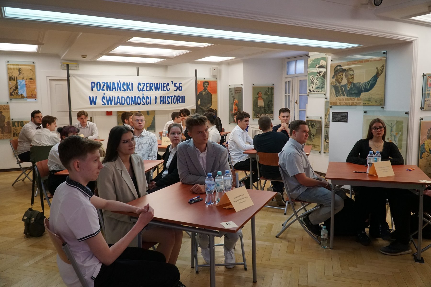 Przy pięciu stolikach siedzą 3-osobowe druzyny uczniów. Są ubrani na galowo. Z tyłu biały transparent z niebieskim napisem 'Poznański Czerwiec '56 w świadomości i historii". Uczniowie siedzą w sali ozdobionej obrazami z okresu PRL-u.