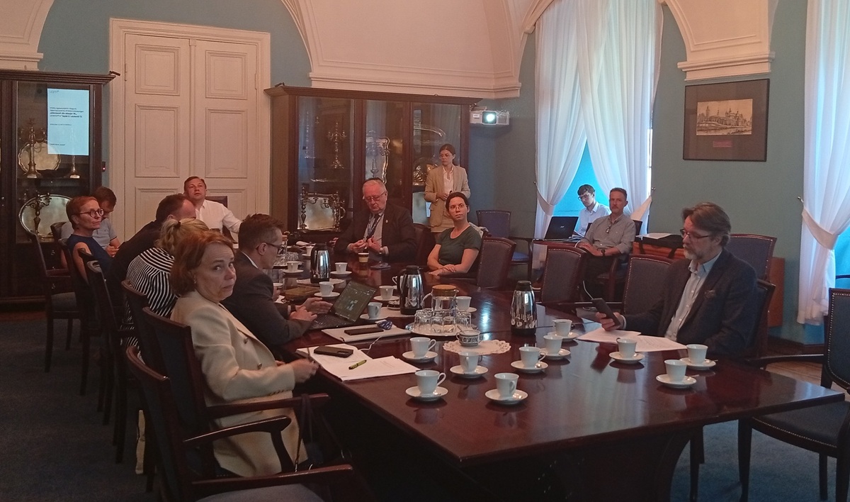 Przy stole konferencyjnym obraduje około kilkanaścioro osób, kobiet i mężczyzn w różnym wieku, patrzą w jednym kierunku, jedna osoba stoi przy projektorze