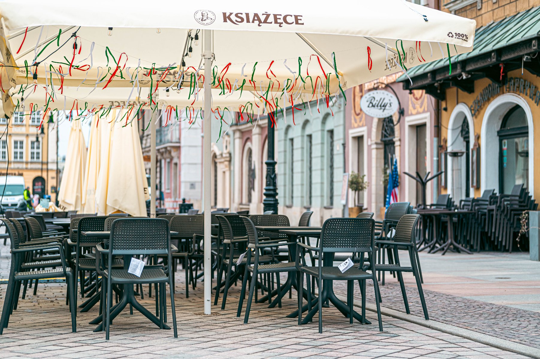 Pusty ogródek gastronomiczny z ciemnoszarymi krzesłami i stolikami ustawionymi na bruku pod dużym, jasnym parasolem z napisem "KSIĄŻĘCE". Brzeg parasola ozdobiony jest wiszącymi, kolorowymi tasiemkami. W tle widoczne są zabytkowe kamienice Starego Rynku, witryny lokali (w tym z szyldem "Billy's") oraz złożone ogródki restauracyjne.