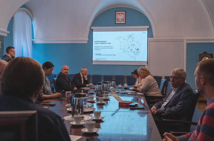 Radni siedzą wzdłuż podłużnego stołu, w tle prezentacja. Referuje ją stojący mężczyzna. - grafika artykułu