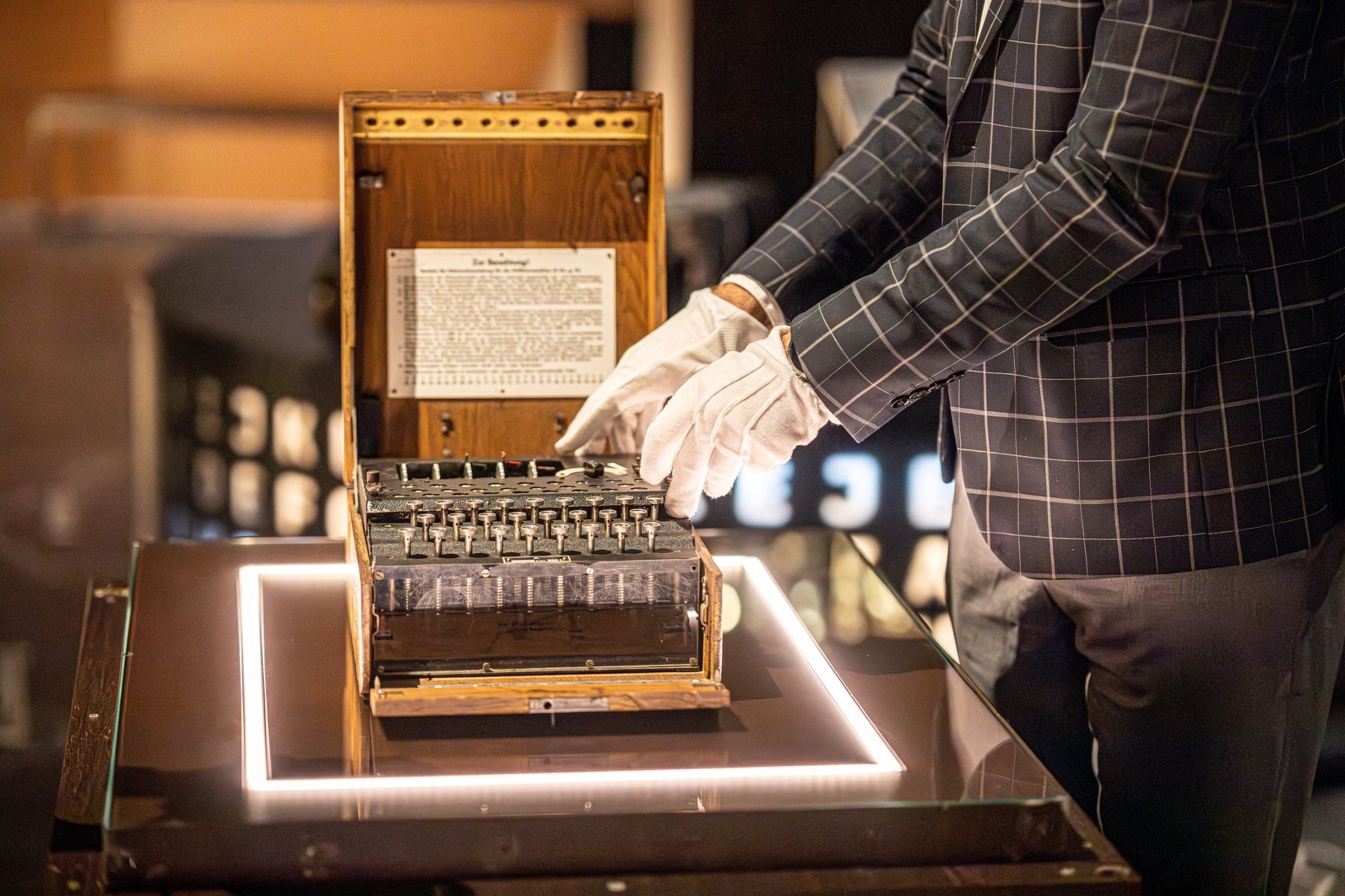 Ręce w białych rękawiczkach, należące do osoby w kraciastej marynarce, dotykają lub prezentują otwartą maszynę szyfrującą Enigma. Maszyna stoi na szklanej podstawce z podświetloną ramką. Wewnątrz widoczne są mechanizmy i klawisze. W tle, rozmyte, znajdują się inne elementy wystawy.