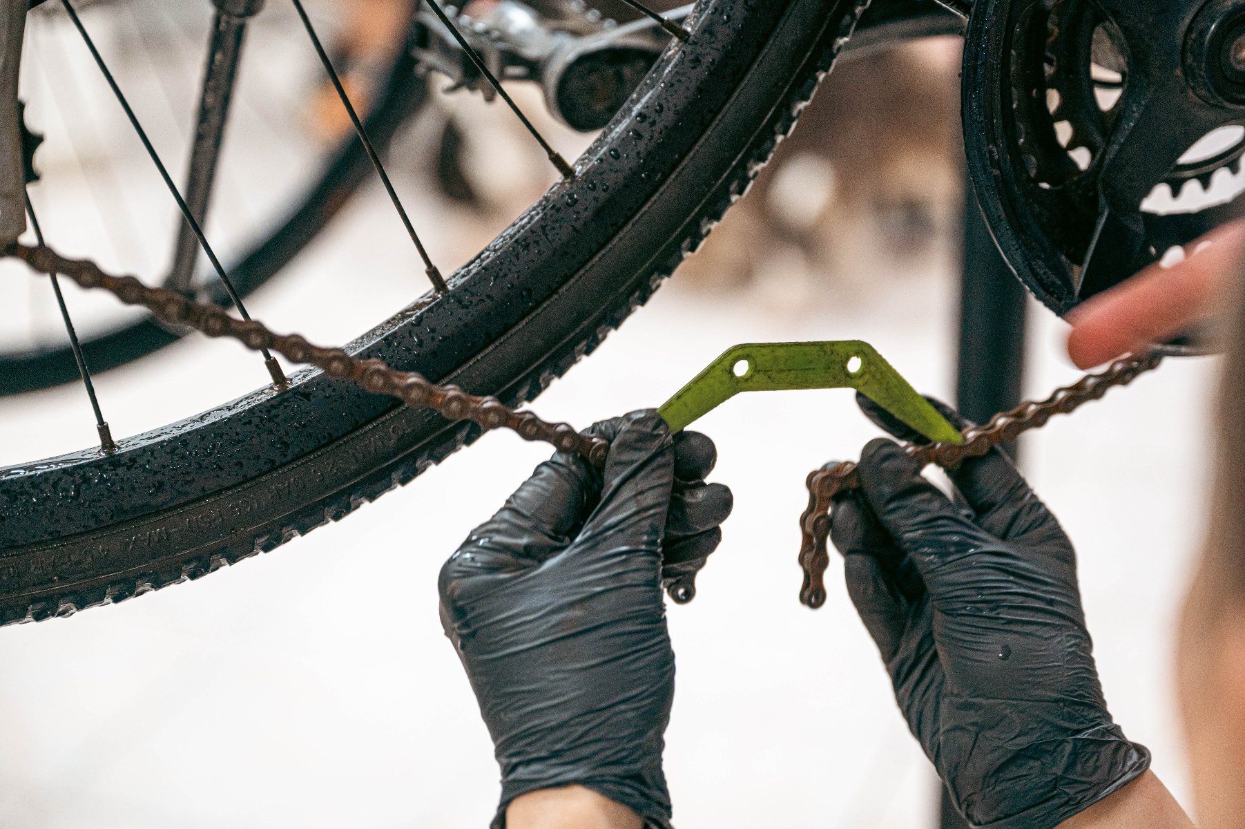 Ręce w czarnych rękawiczkach trzymają łańcuch rowerowy i zielony miernik zużycia łańcucha, sprawdzając jego stan. W tle widoczne jest koło roweru i zębatka.