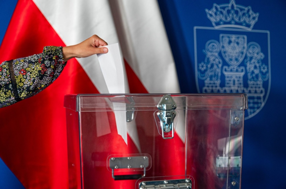 ręka wrzuca kartę do przezroczystej urny wyborczej, w tle flaga Polski o herb Poznania