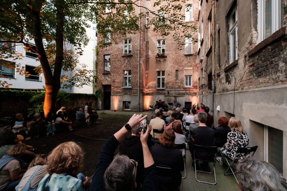 Robi się ciemno, artyści kończą występ i kłaniają się publiczności, która siedzi na rozłożonych na podwórzu krzesłach.