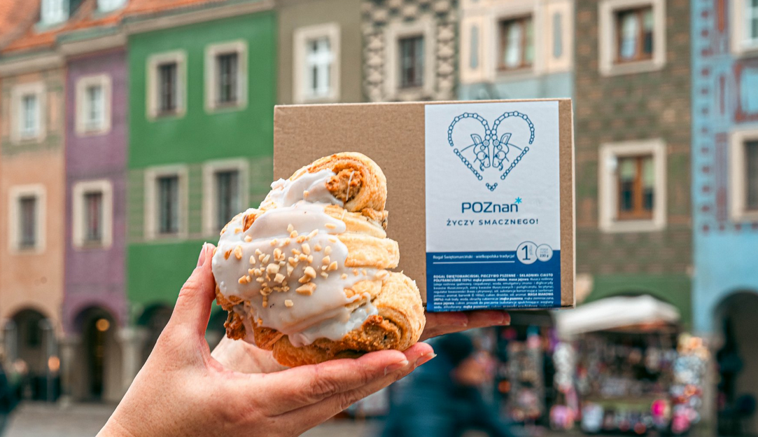Rogal świętomarciński na tle kamieniczek na Starym Rynku. Za rogalem kartonik a na nim napis Poznań życzy smacznego.