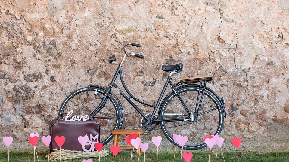 Rower oparty o starą ścianę, przed rowerem wbite w ziemię patyczki z papierowymi serduszkami, obok roweru teczka z napisem "love".