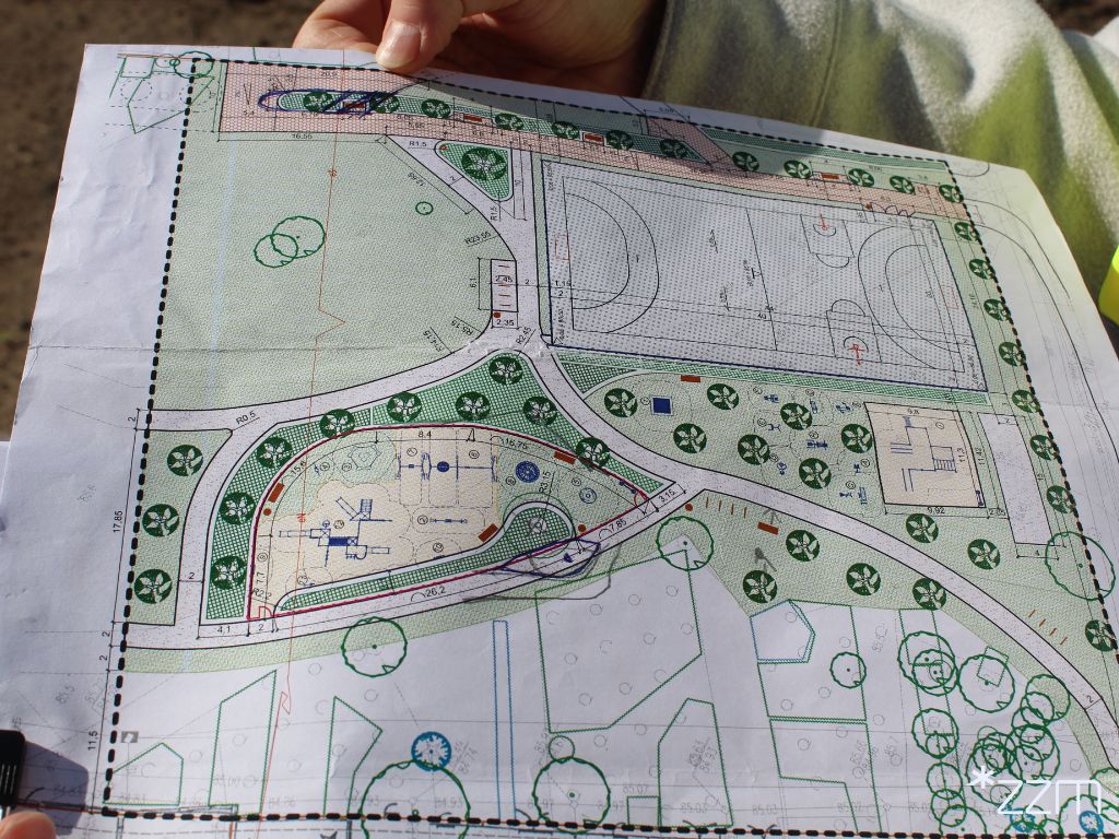 Rzut z góry przedstawiający fragment architektonicznego planu zagospodarowania terenu. Na projekcie widoczny jest plac zabaw, boisko sportowe oraz ścieżki i tereny zielone z symbolami drzew. Plan jest wydrukowany na kartce, którą trzymają dwie dłonie.