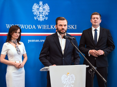 rzy osoby w formalnych strojach stoją na tle niebieskiego baneru z napisem "WOJEWODA WIELKOPOLSKI". Mężczyzna w centrum przemawia do mikrofonu, a kobieta w białej sukience stoi po jego lewej stronie. Na mównicy znajduje się godło.