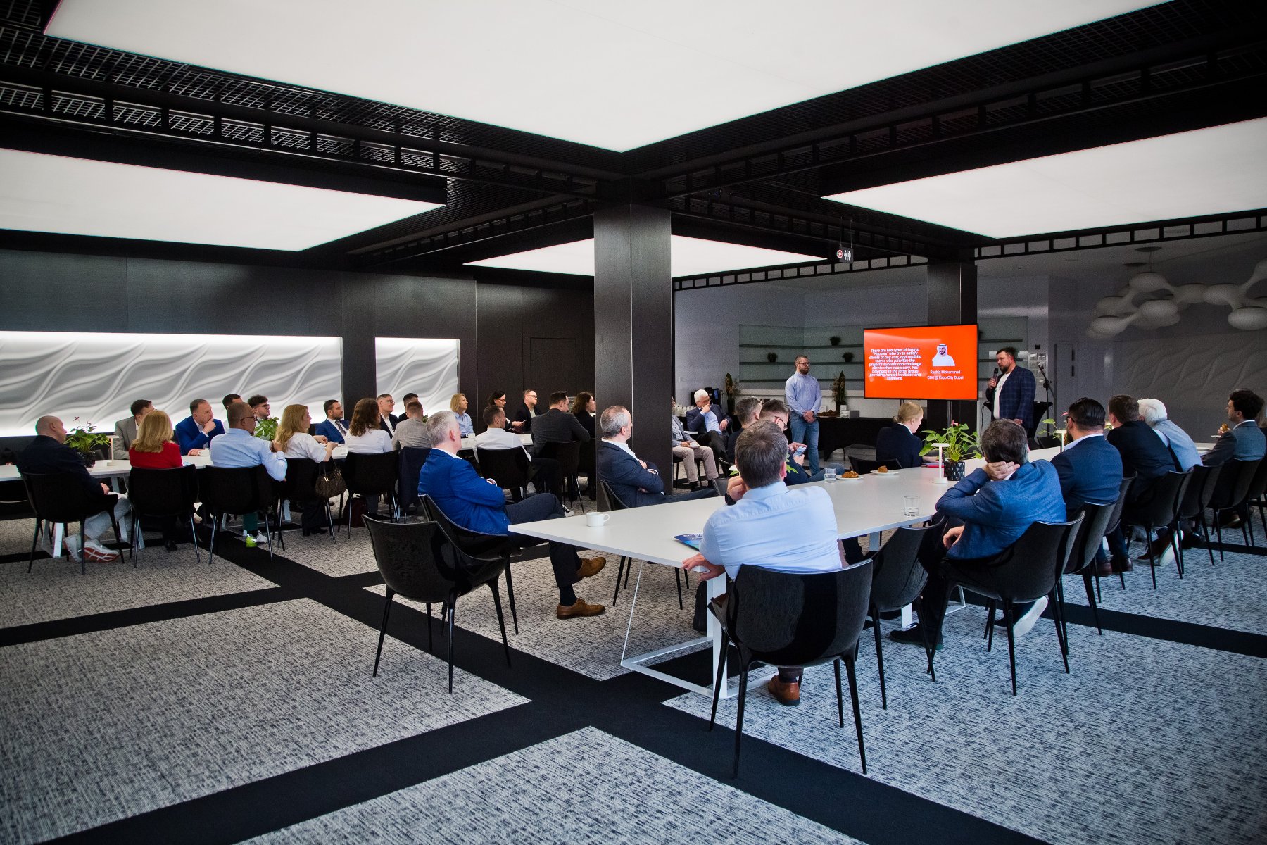 Sala konferencyjna, przy stołach siedzą ludzie, przed nimi ekran, na której wyświetlana jest prezentacja.
