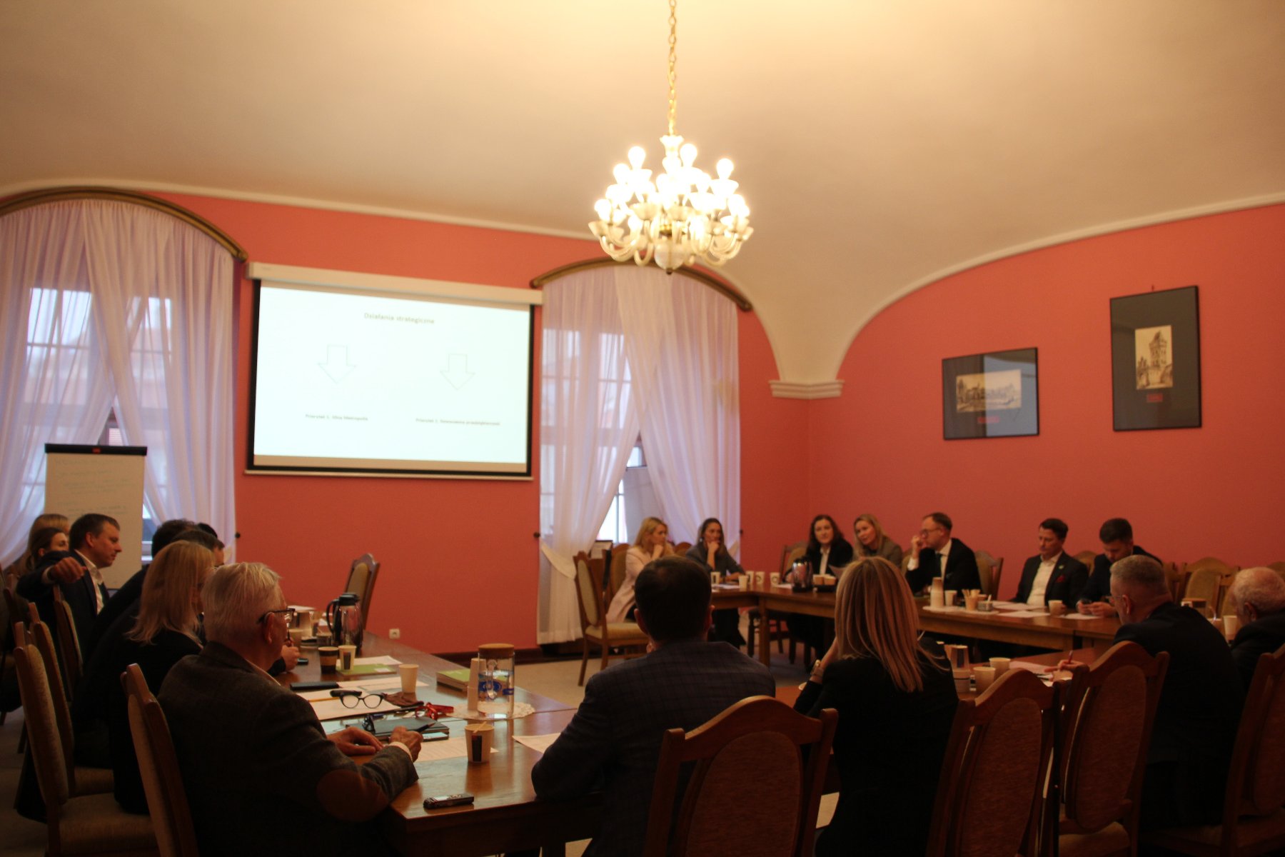 Sala Malinowa. Przy stole w kształcie podkowy siedzą kobiety i mężczyźni w eleganckich strojach i prowadzą rozmowy.