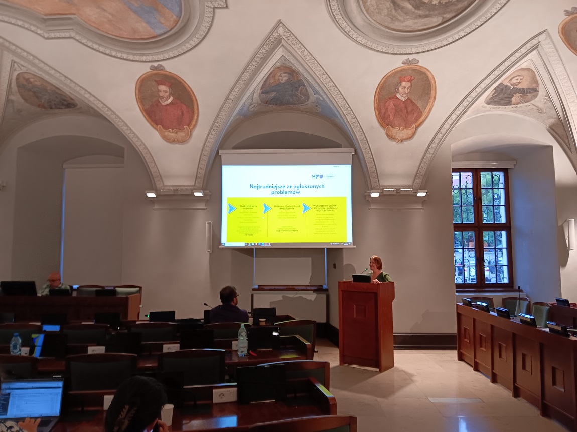 sala obrad z sufitem zdobionym kolorowymi freskami, jedna osoba stoi przy mównicy, na ekranie wyświetlona jest prezentacje, pozostałe osoby siedzą i słuchają