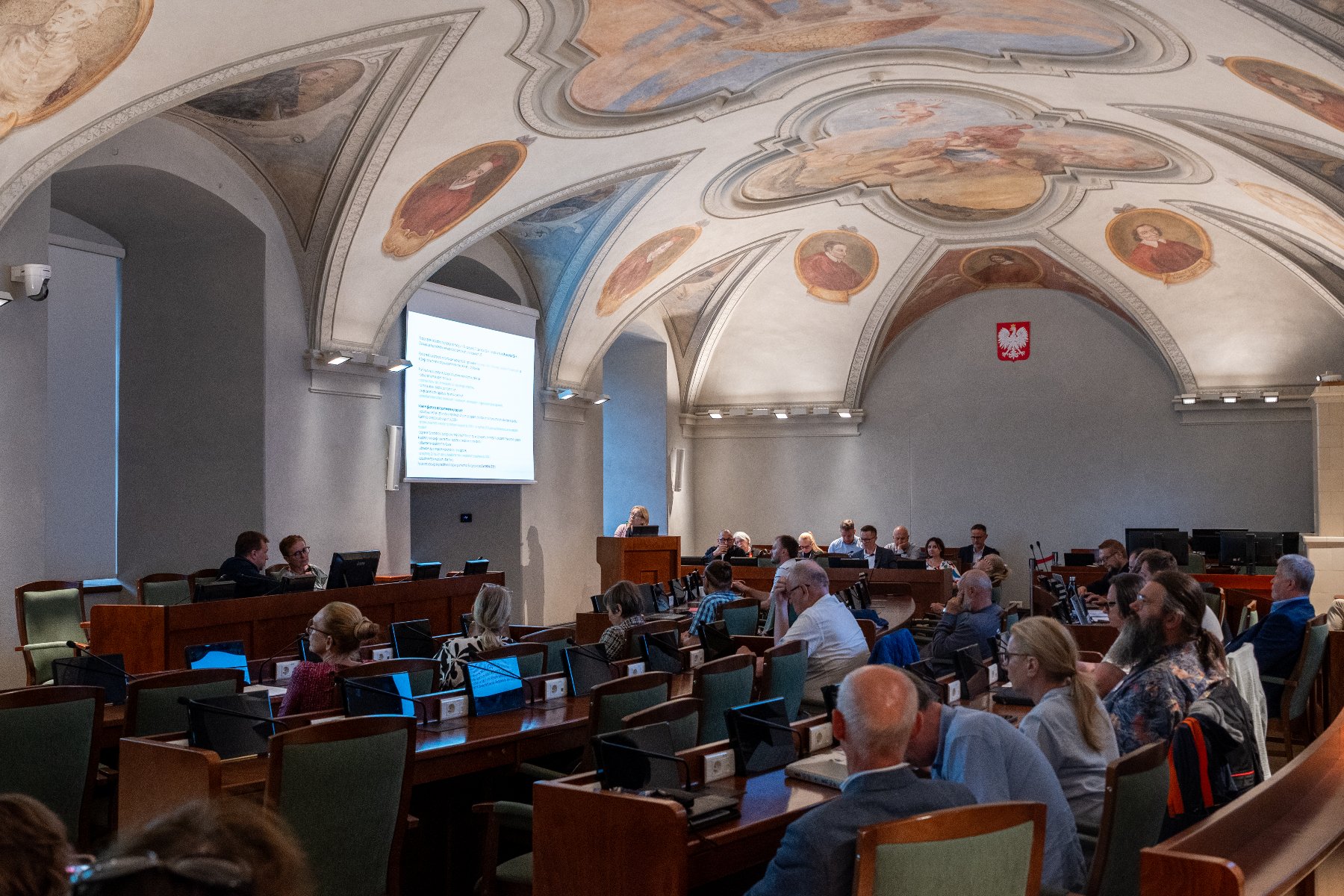 Sala sesyjna. Radni i mieszkańcy siedzą przy stołach.