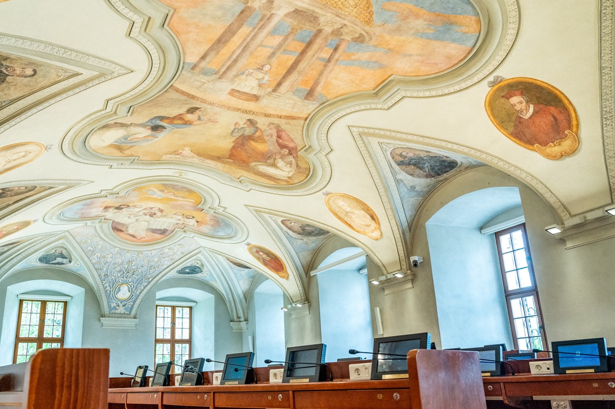 sala sesyjna Rady Miasta Poznania widać sufit zdobiony barwnymi zabytkowymi freskami oraz stanowiska dla radnych z monitorami i mikrofonami