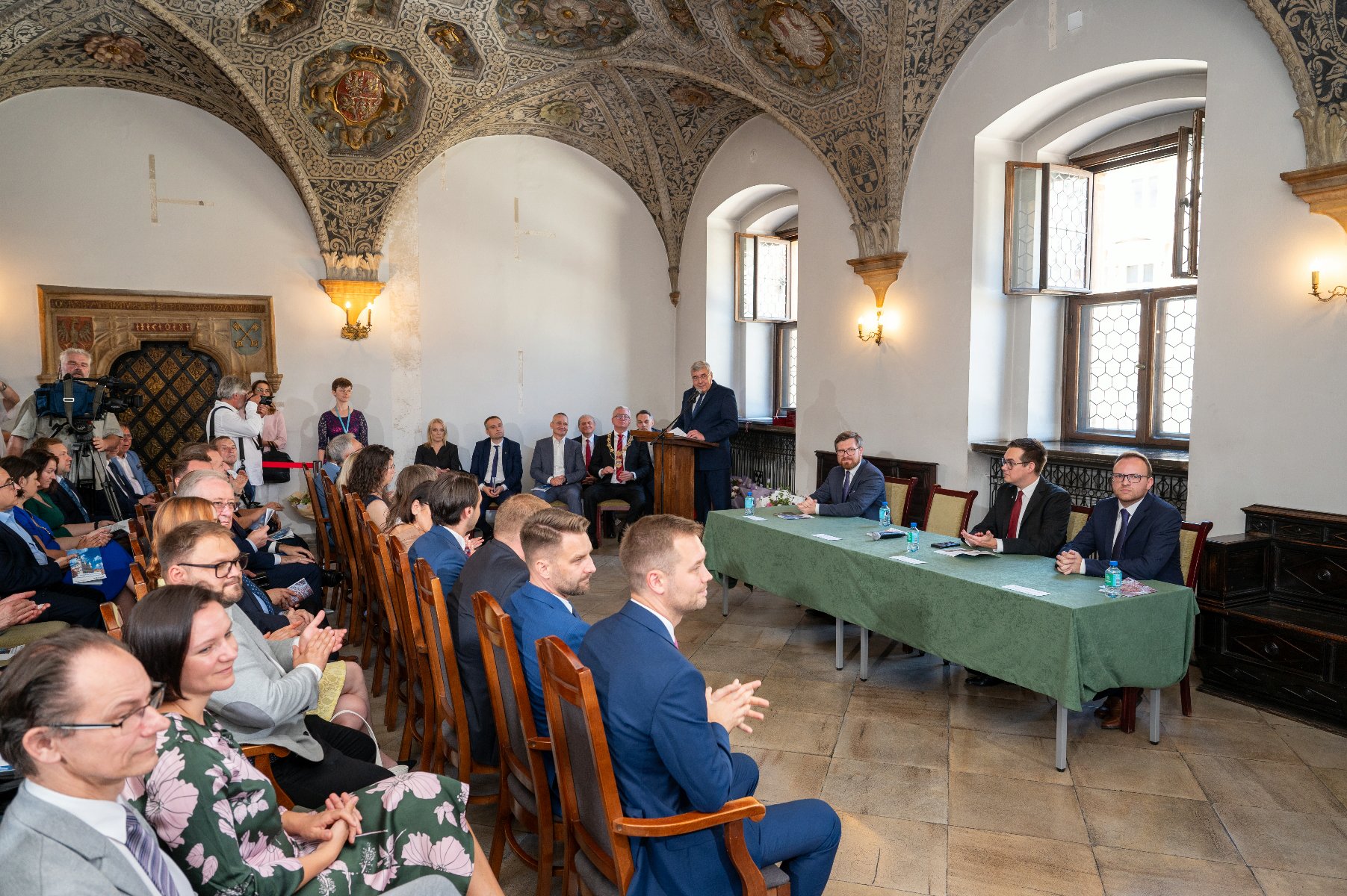 Sala z łukowymi sklepieniami i bogato zdobionym sufitem. Na pierwszym planie widoczni są goście siedzący na drewnianych krzesłach. Po prawej stronie, za stołem nakrytym zielonym obrusem, siedzą czterej mężczyźni. W tle, przy mównicy, stoi mężczyzna i przemawia.
