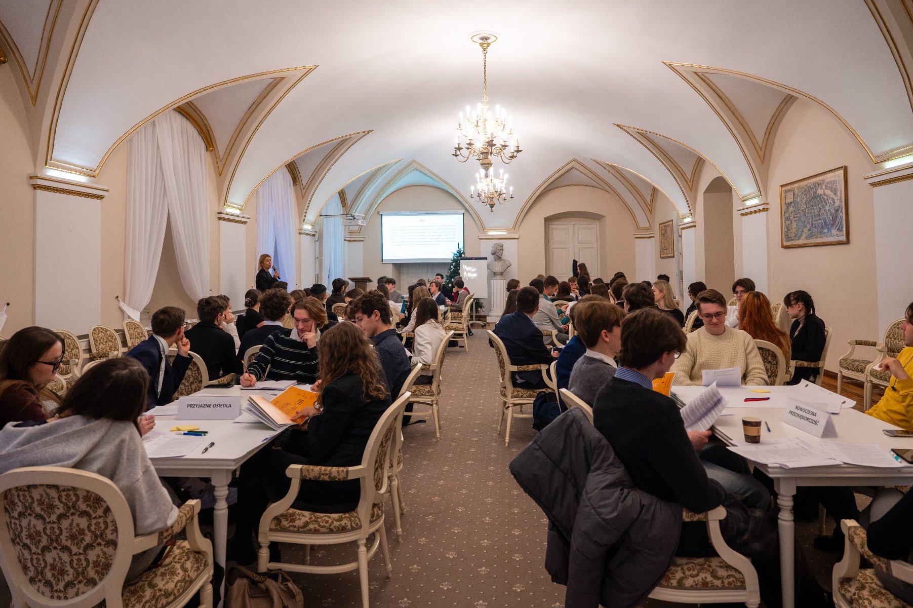 Sala z ozdobnymi krzesłami i złotymi zdobieniami na ścianach przy stolach siedzą grupki młodzieży pracującej w grupach