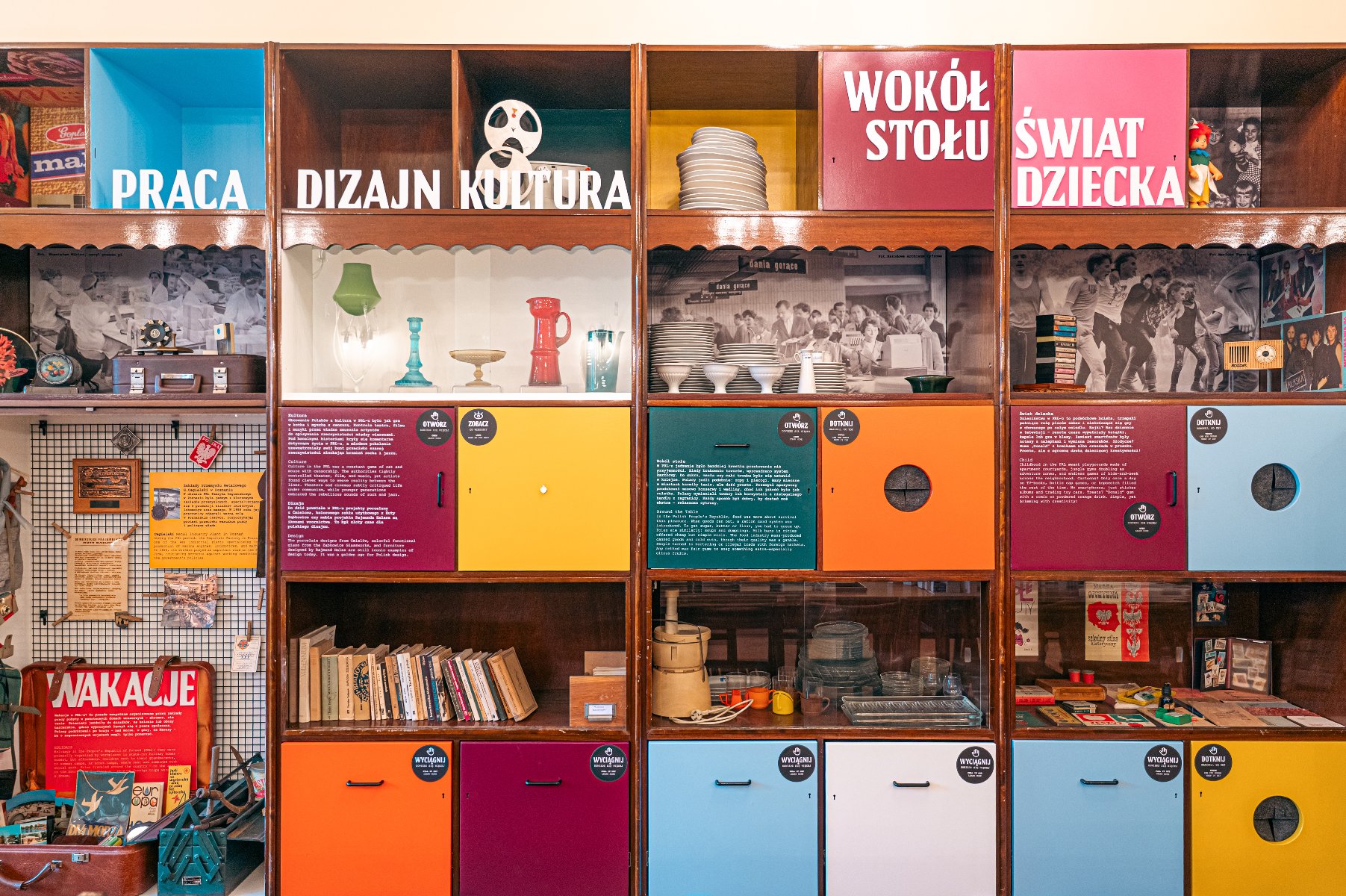 Salon w stylu vintage, z różnymi retro elementami. Pokój ma tapetę w czerwone kwiatowe wzory, drewnianą szafkę z półkami, na których znajdują się różne przedmioty, oraz oprawiony obraz Jezusa na ścianie. Na drewnianym stojaku stoi telewizor w stylu retro, pod którym leży koronkowa serwetka. Obok telewizora znajduje się lampka podłogowa z żółtym abażurem. Przed telewizorem stoi drewniany stolik kawowy z gazetami, szklana popielniczka i kilka innych przedmiotów.