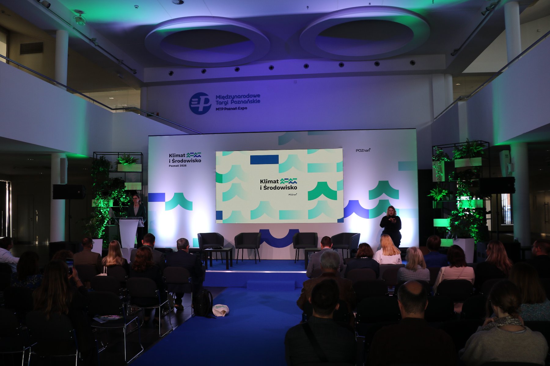 Scena, na której znajduje się baner z logo konferencji. W tle widać logotyp Międzynarodowych Targów Poznańskich. Na scenie znajduje się kobieta przy pulpicie mówniczym. Z drugiej strony tłumacz języka migowego.